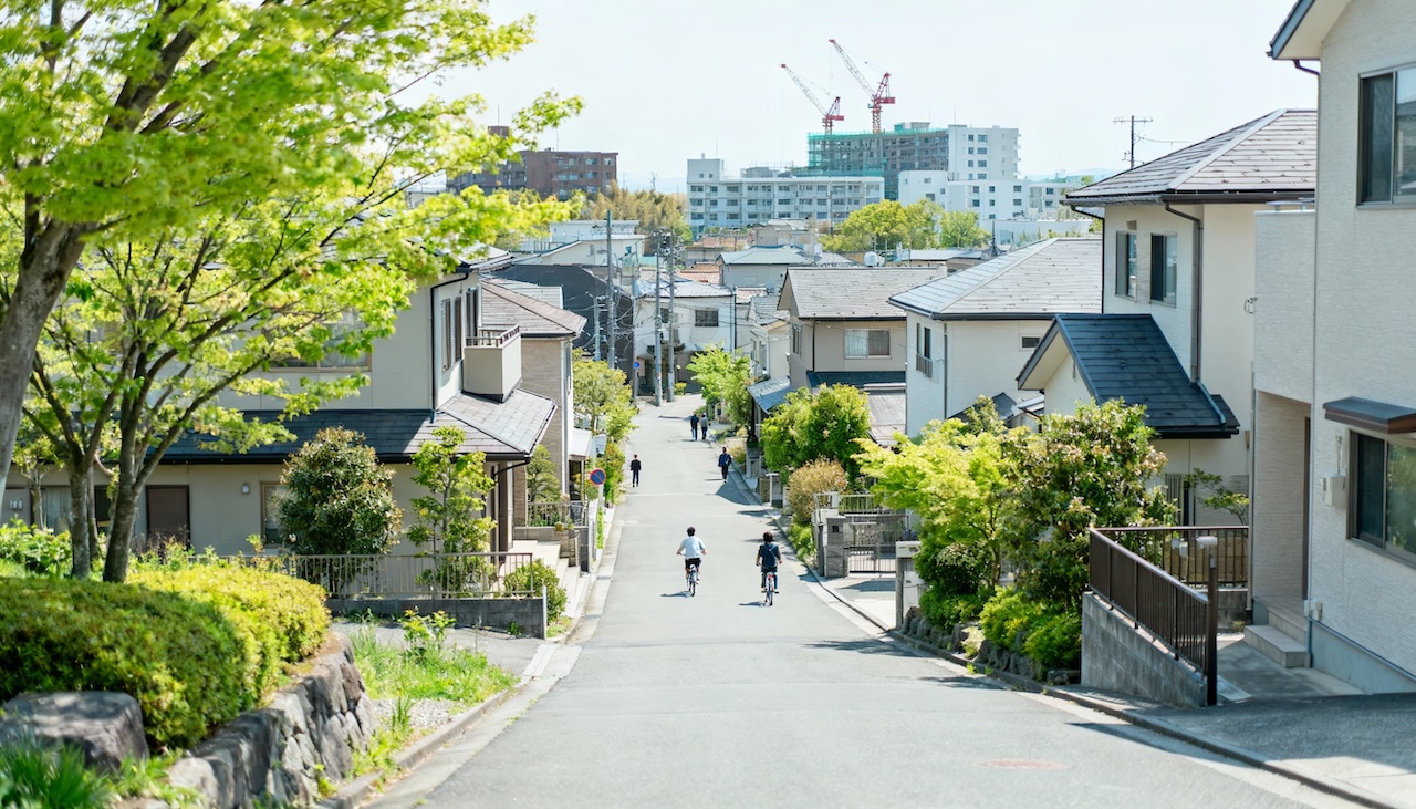 緑に囲まれた住宅街の坂道を人々が行き交う風景